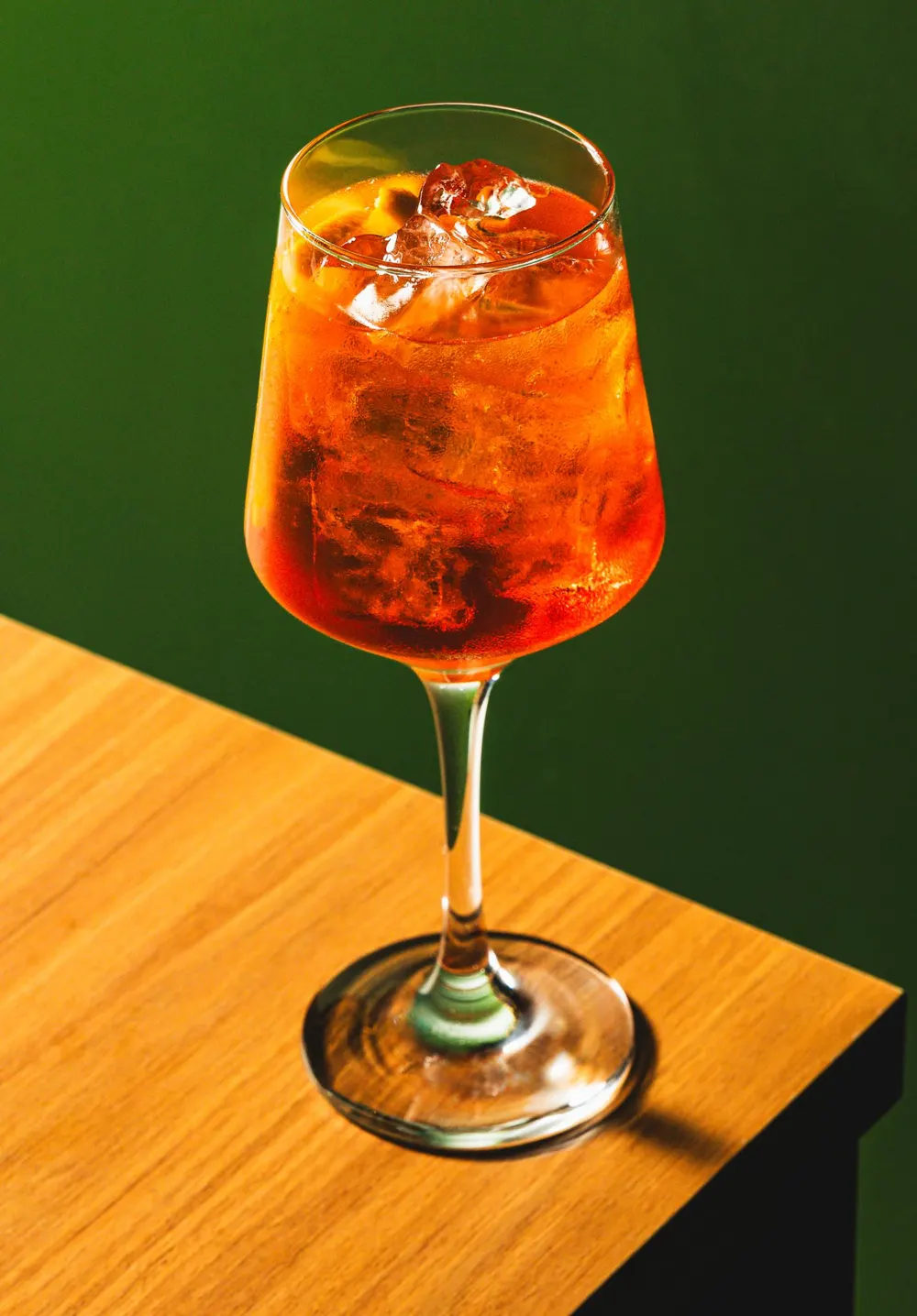 Aperol Spritz em taça sobre mesa de madeira, fundo verde